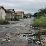 Rua deteriorada em Nova Colina, Brasília, com buracos, lixo e prédios abandonados, expondo descaso e falhas graves.