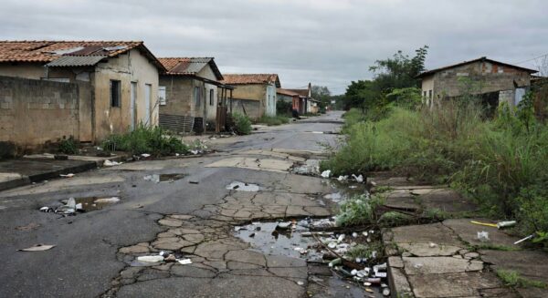 Rua deteriorada em Nova Colina, Brasília, com buracos, lixo e prédios abandonados, expondo descaso e falhas graves.