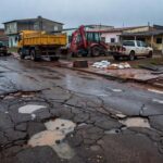 Rua danificada por chuvas no P Sul de Ceilândia, com equipamentos de reparo e veículos de manutenção ao fundo.
