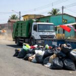Rua em Ceilândia com mutirão de limpeza, sacos de lixo e caminhões coletando resíduos para celebrar 55 anos e combater pragas.